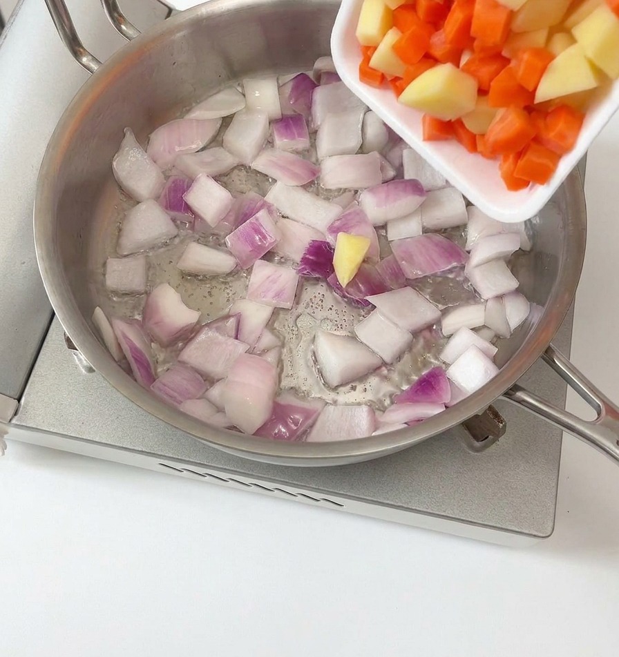 Patate e carote a cubetti versate da un vassoio bianco in una padella dacciaio contenente cipolle rosse soffritte.