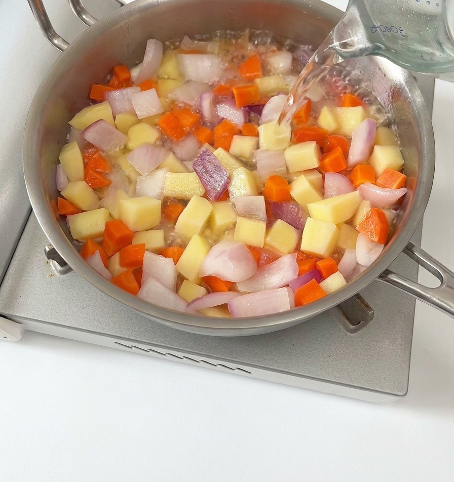 Acqua versata da un misurino di vetro in una padella piena di carote, patate e cipolle rosse a dadini.