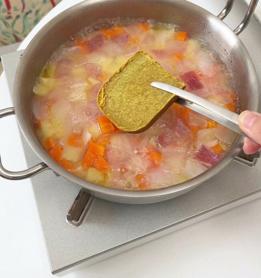 Pinze di metallo che tengono un dado di roux per curry marrone sopra una padella fumante di verdure e acqua.