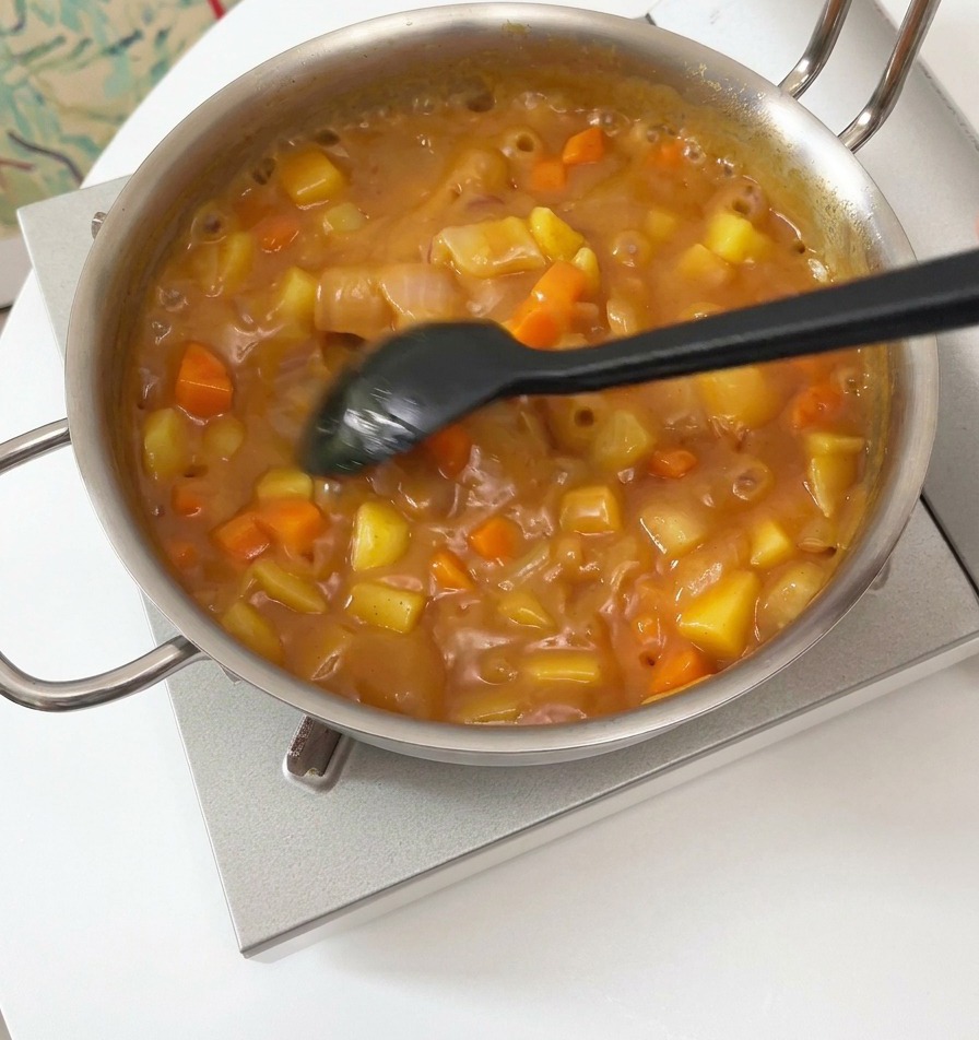 Una spatola nera che mescola una salsa curry densa e gorgogliante con pezzi visibili di patate e carote in una pentola argentata.