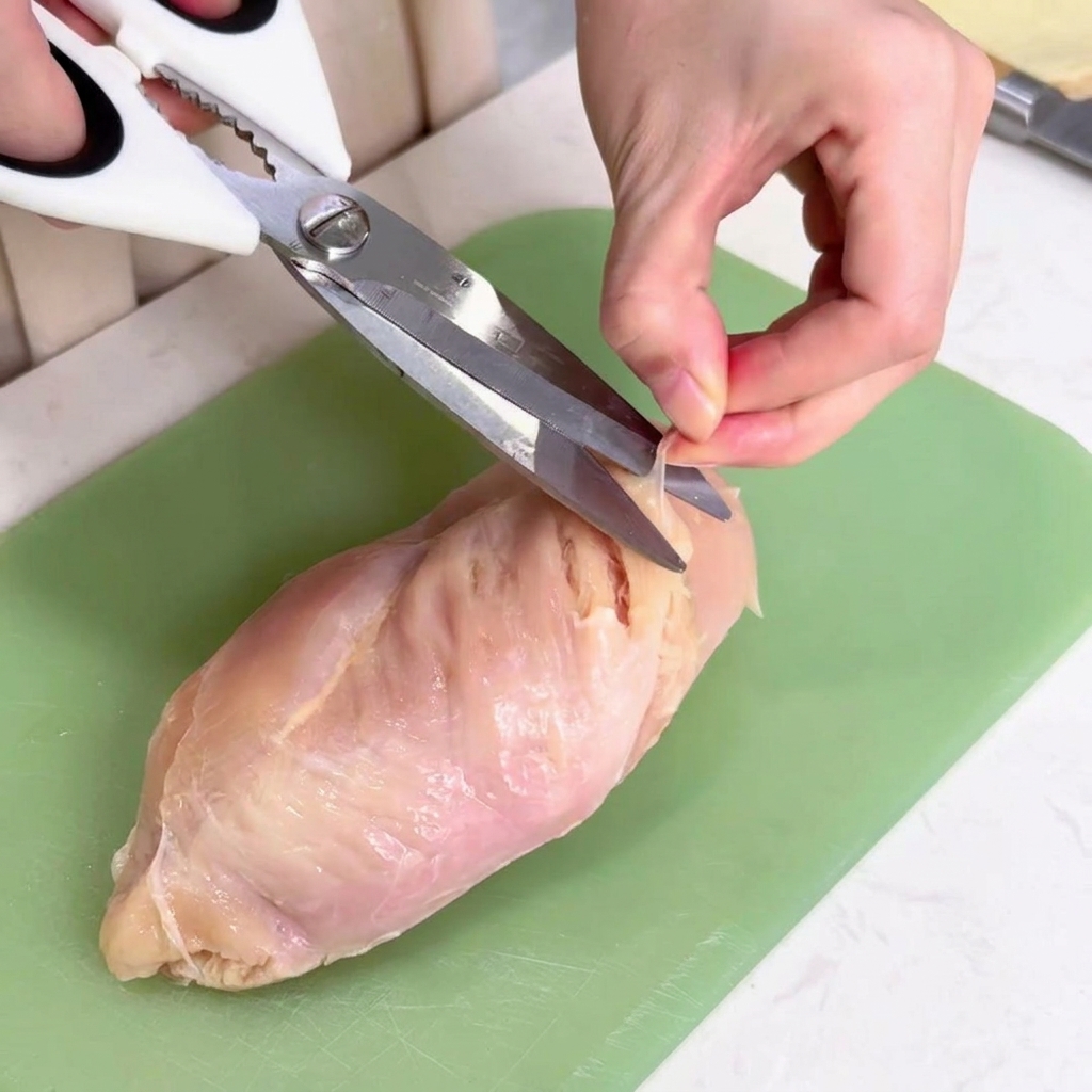 Una mano che usa forbici da cucina per tagliare via la sottile membrana bianca dal petto di pollo su un tagliere verde.
