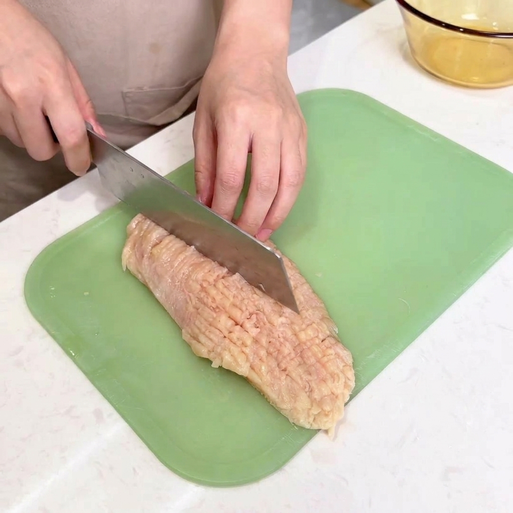Il dorso di un coltello da chef usato per battere e intenerire un pezzo di pollo piatto su un tagliere.