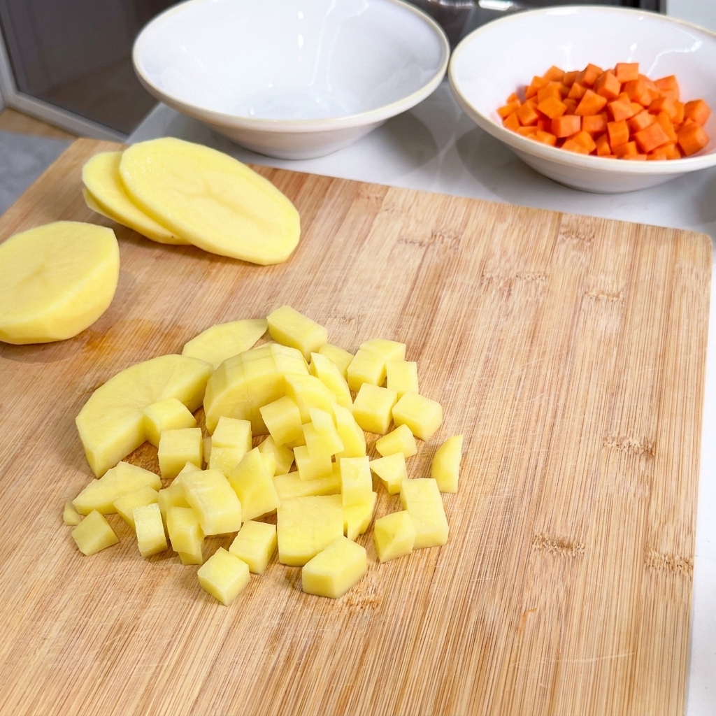 Cubetti di patata gialla ammucchiati su un tagliere accanto a una ciotola di carote già tagliate.
