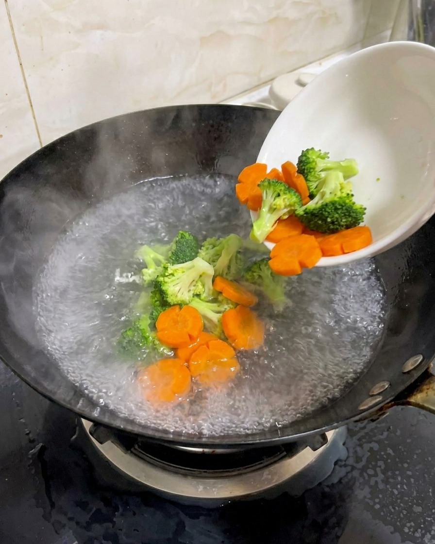 Broccoli freschi e carote a forma di fiore versati da una ciotola bianca in un wok con acqua bollente.