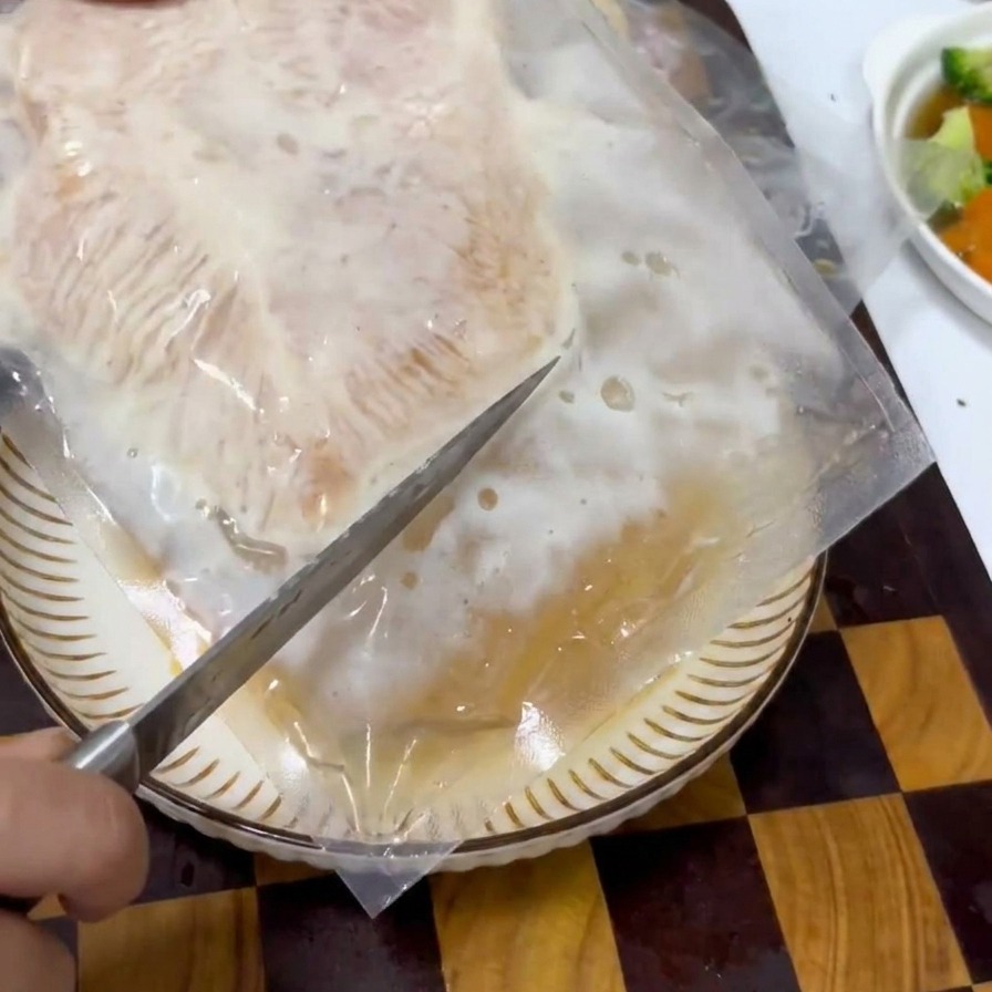 Una mano che usa un coltello per aprire una confezione sottovuoto contenente una fetta di pollo marinato.