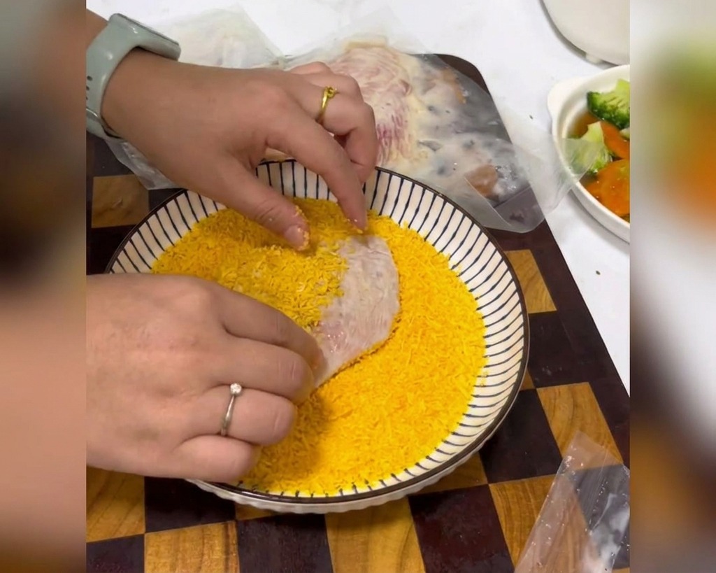 Mani che premono un petto di pollo marinato in una ciotola bianca piena di pangrattato giallo brillante.
