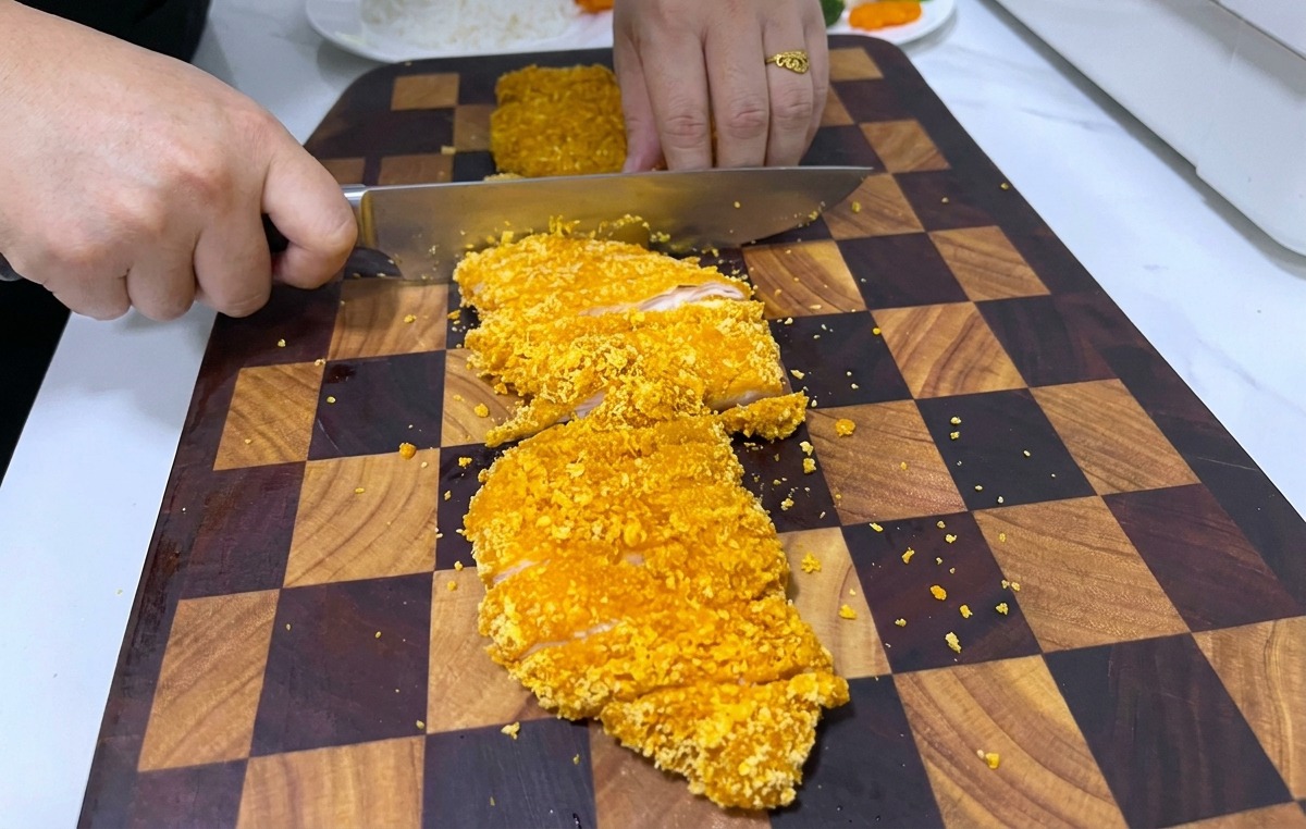 Una persona che usa un grosso coltello per affettare una cotoletta di pollo croccante su un tagliere di legno.