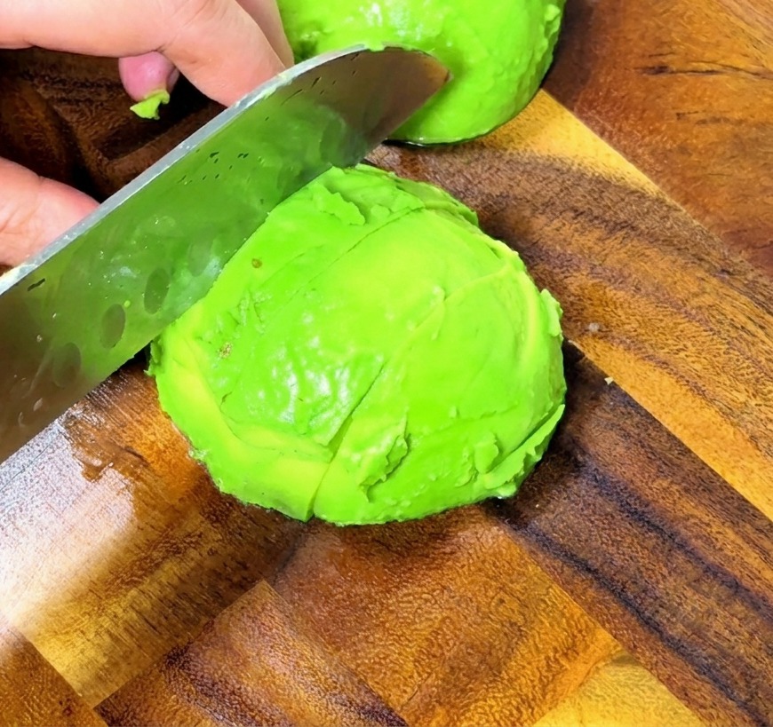 Primo piano di mani che tagliano un avocado sbucciato a cubetti uniformi su un tagliere di legno.