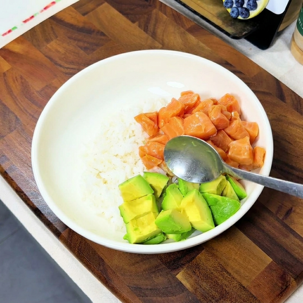 Cubetti di avocado aggiunti in una ciotola che contiene già riso e salmone marinato.