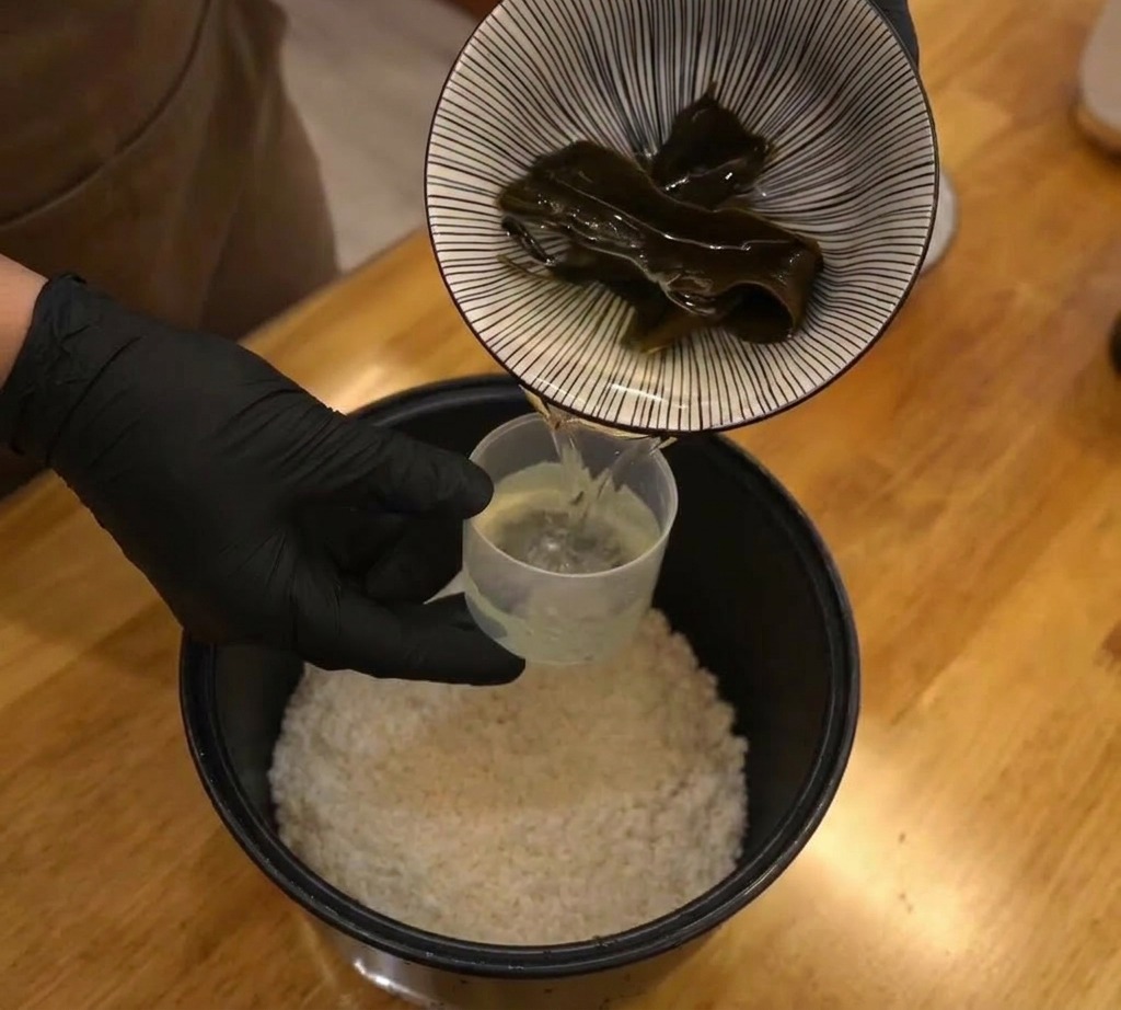 Un pezzo di alga kombu essiccata che viene inserito in una pentola per riso con riso bianco e acqua.