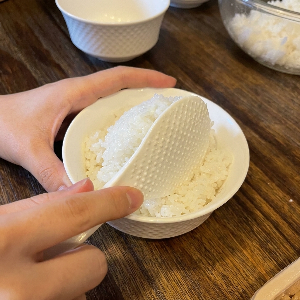 Una spatola per riso che depone una porzione arrotondata di riso condito in una ciotola di ceramica bianca.