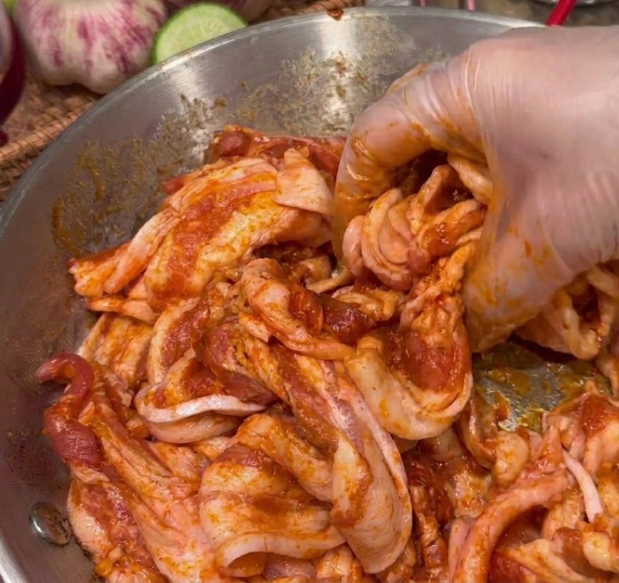 Una mano che mescola fette di pancetta di maiale con una marinatura densa e rossa in una ciotola di acciaio inossidabile.