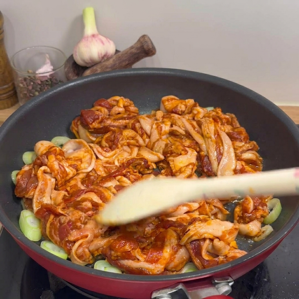 Primo piano della pancetta di maiale che sfrigola in padella con cipollotti, girata con una spatola di legno.