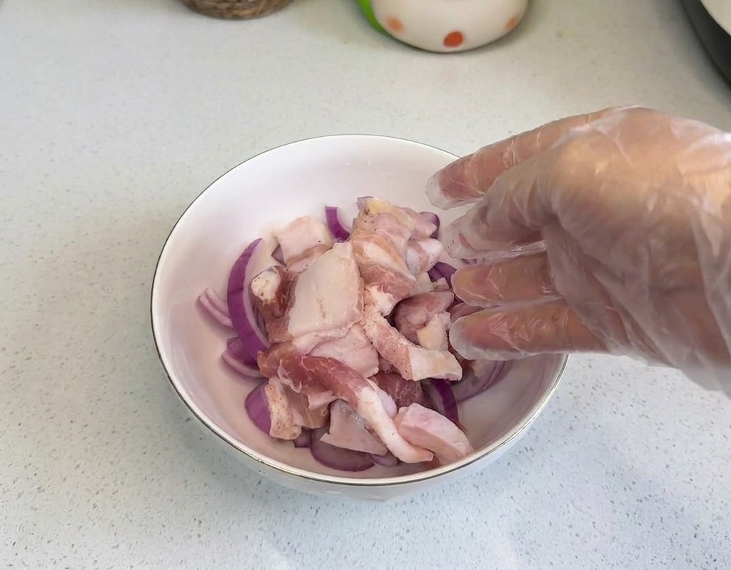 Una mano con un guanto di plastica trasparente che adagia fette di pancetta cruda in una ciotola con anelli di cipolla.