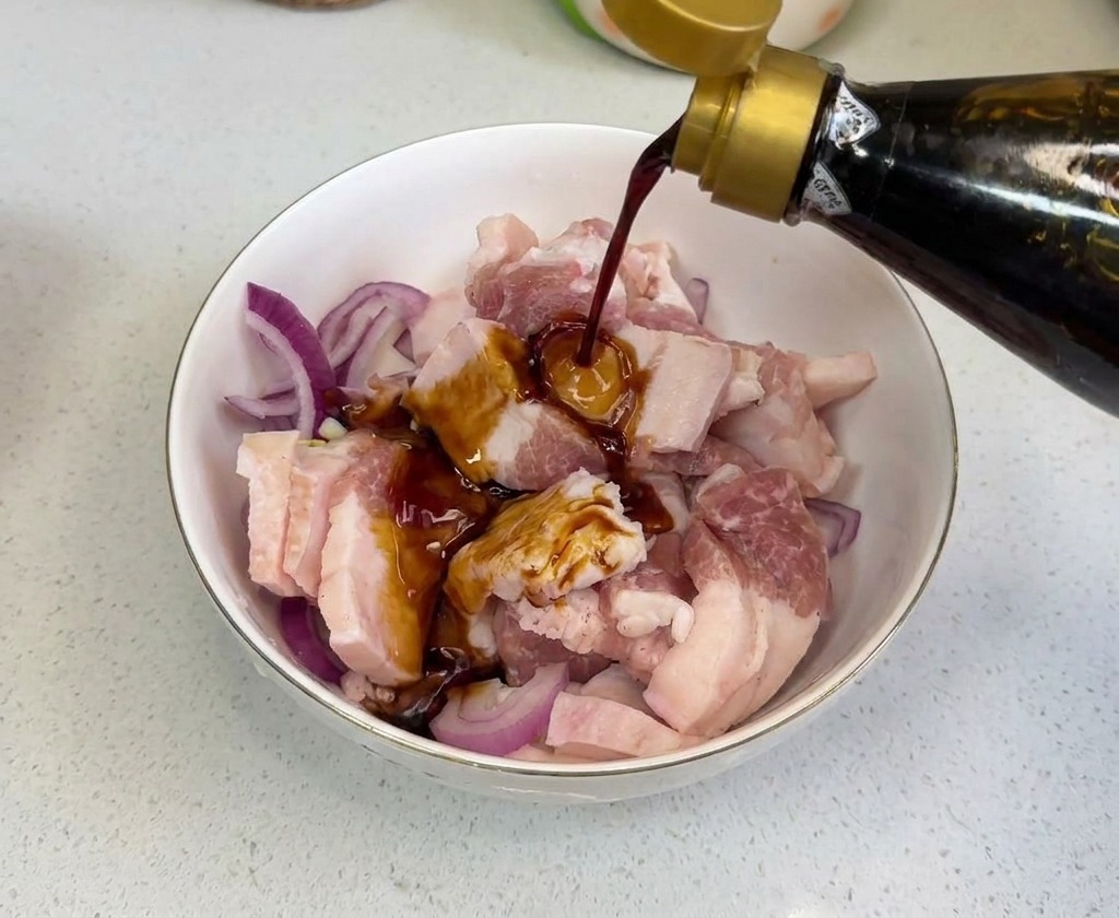 Salsa di soia scura versata da una bottiglia di vetro sulla carne cruda e sulle cipolle in una ciotola.