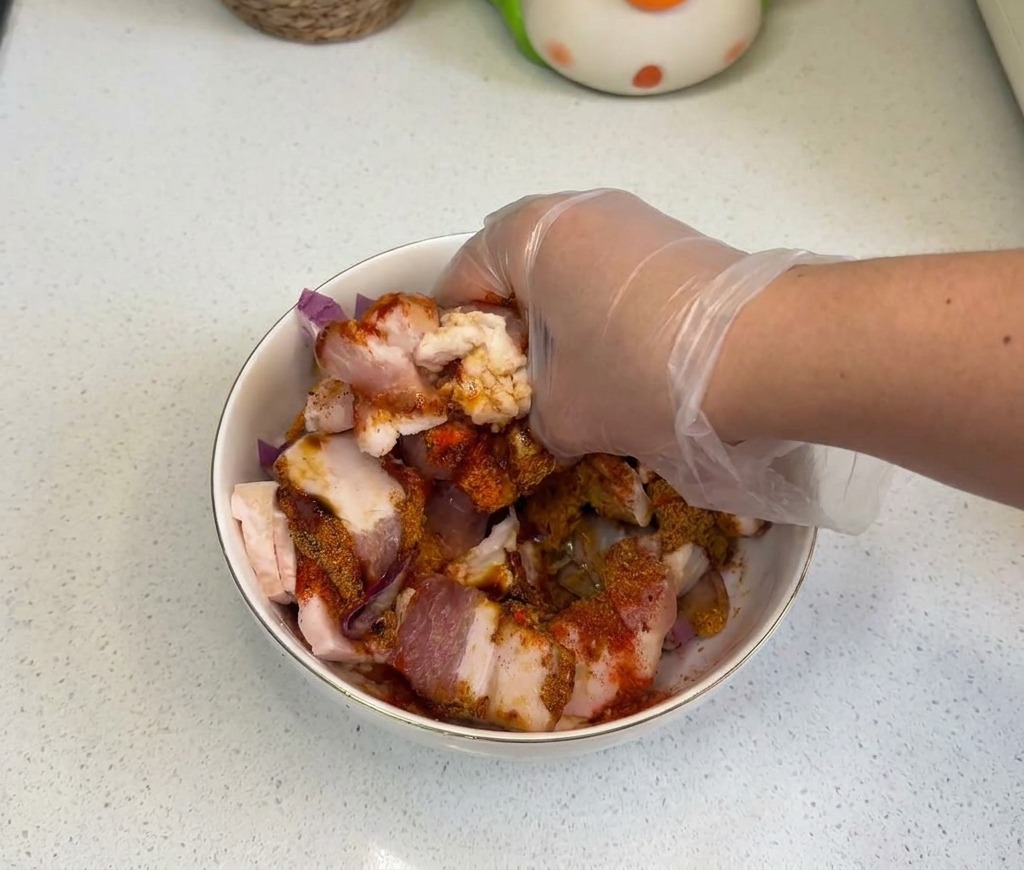 Una mano con guanto che mescola accuratamente pancetta cruda, cipolle e aromi in una ciotola bianca.