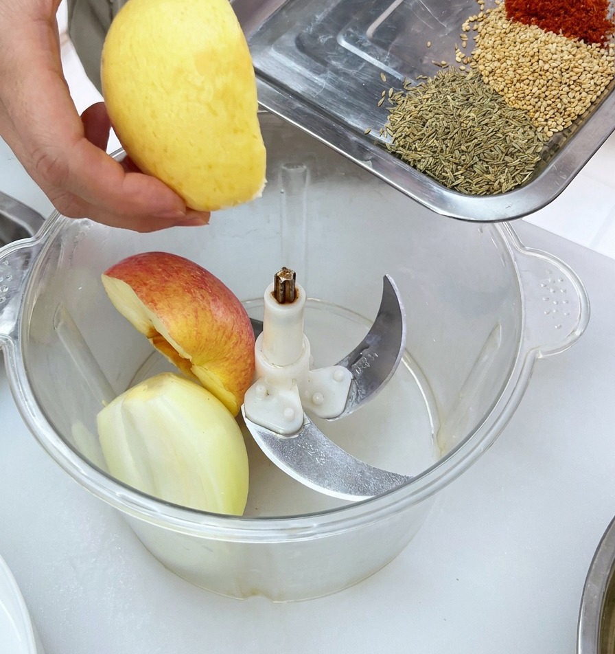 Pezzi di frutta fresca inseriti in un robot da cucina, con un vassoio di spezie pronto per essere aggiunto.
