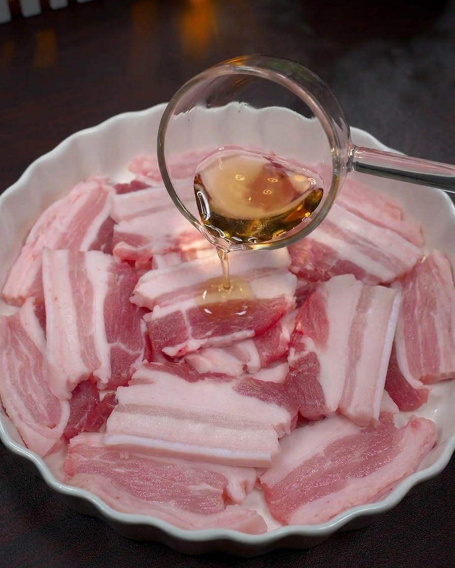 Un liquido trasparente viene versato con un mestolo di vetro sulle fette di carne cruda in una ciotola bianca.