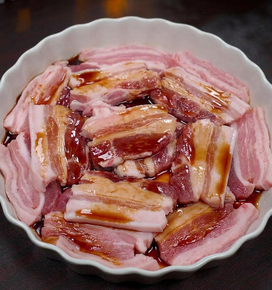 Fette di pancetta cruda in una pirofila bianca mentre vengono bagnate con una marinata scura.