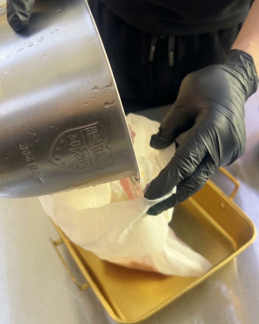 Mani con guanti che strizzano un sacchetto di tela bianca per far gocciolare il succo in una teglia dorata.