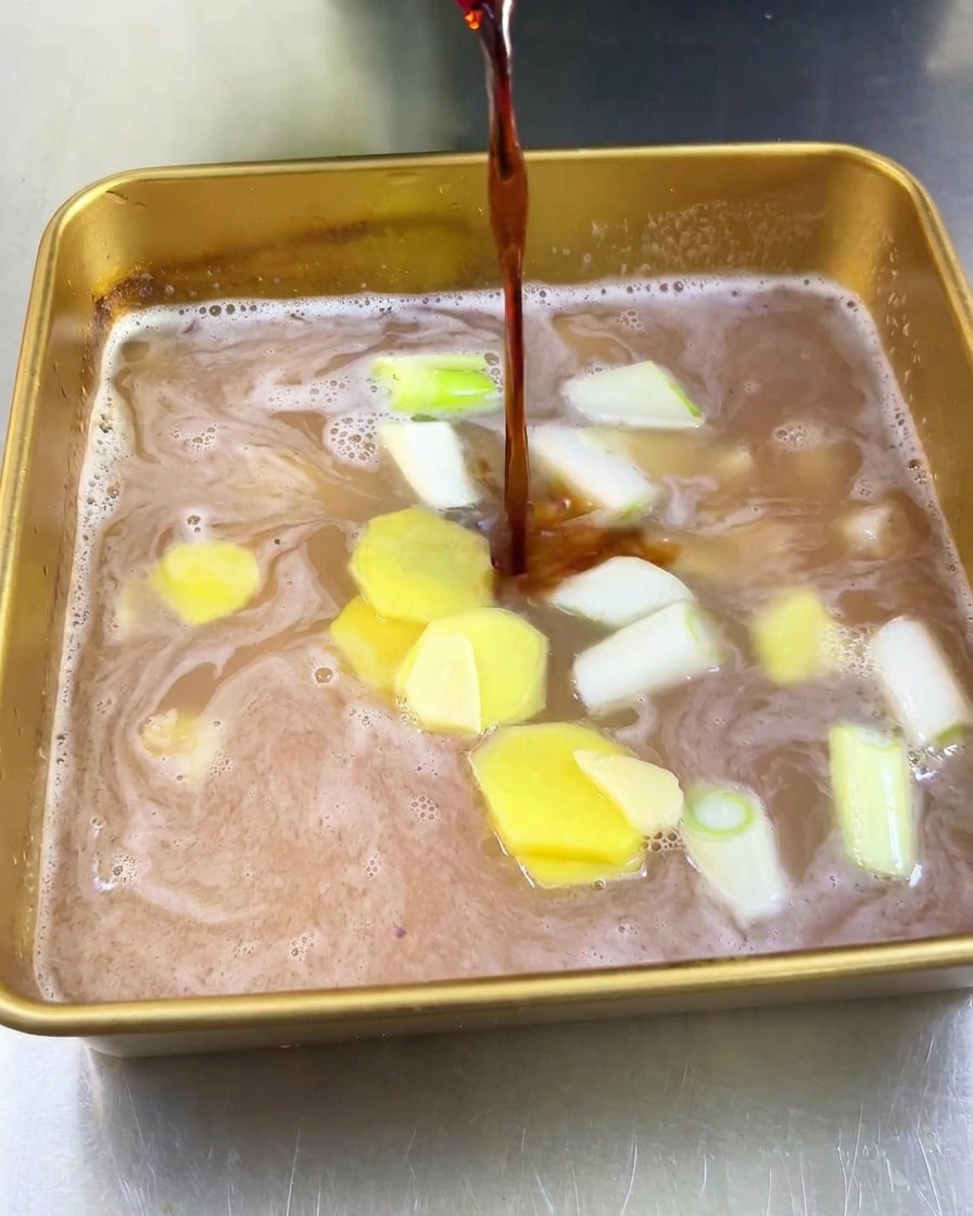Un filo di salsa di soia scura versato in una teglia con succo di frutta, zenzero e cipollotti.