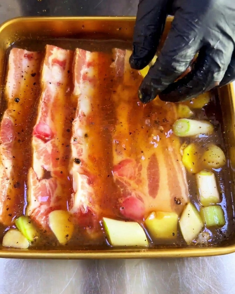 Una mano con guanto che preme fette spesse di pancetta cruda in una teglia di marinata scura.