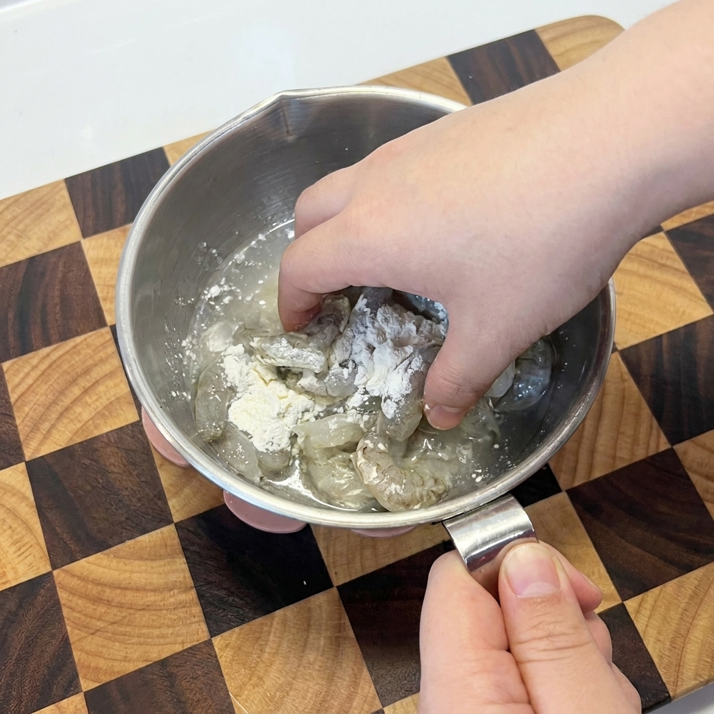 Gamberi crudi mescolati con amido di mais bianco a mano in una ciotola di metallo.