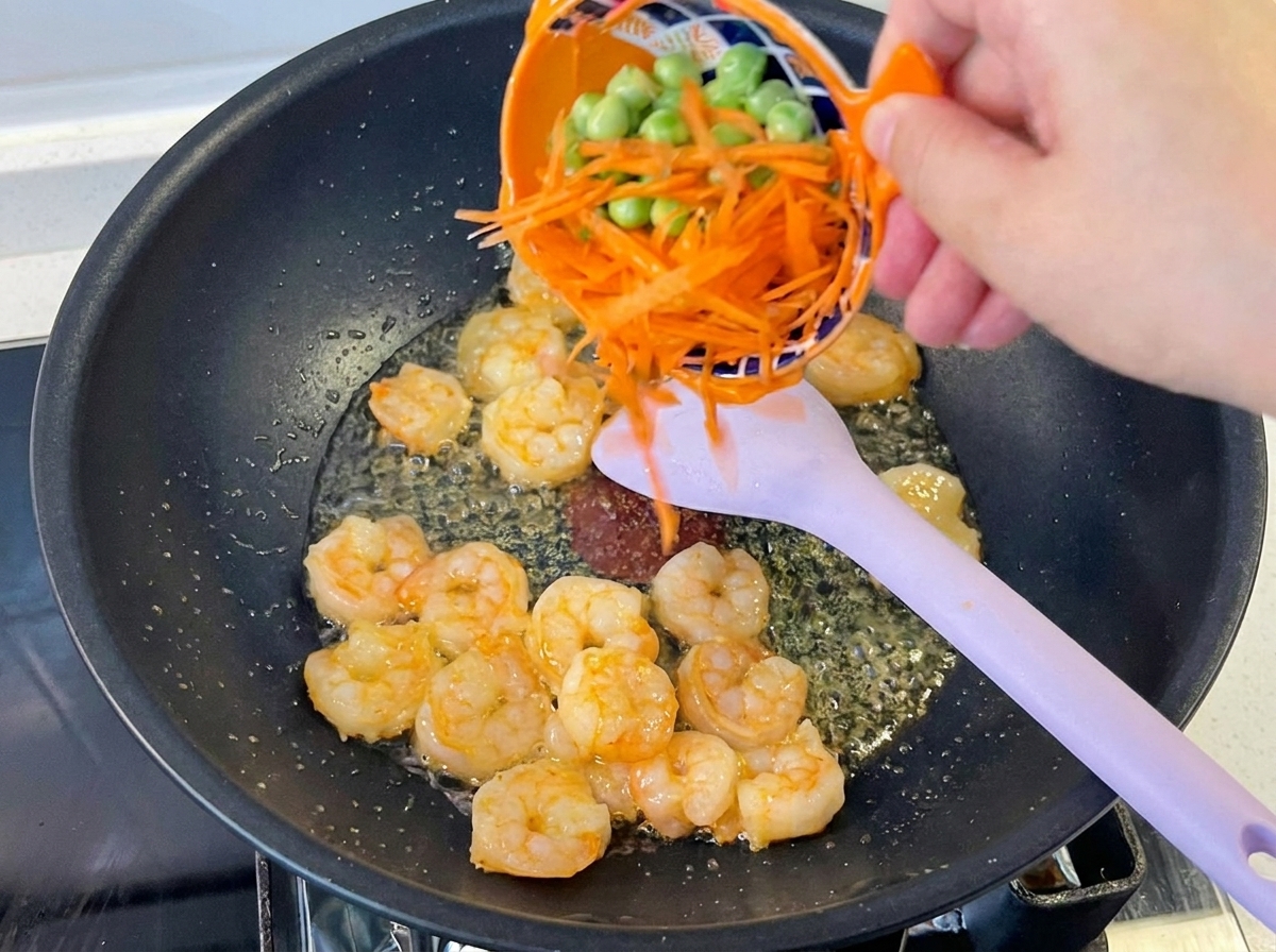 Una mano versa carote a listarelle e piselli verdi in un wok con gamberi già cotti.
