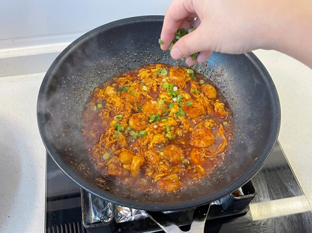 Una mano cosparge cipollotto fresco tritato su un piatto di gamberi e uova fumanti in padella.