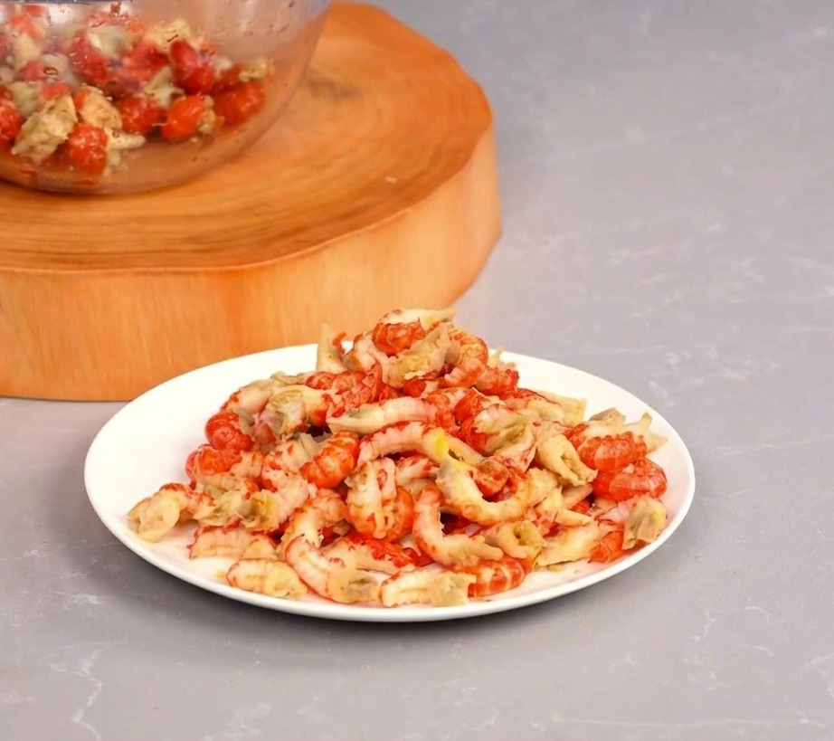 Un piatto bianco colmo di code di gambero di fiume rosse e sgusciate, accanto a una ciotola di vetro su un tagliere di legno.