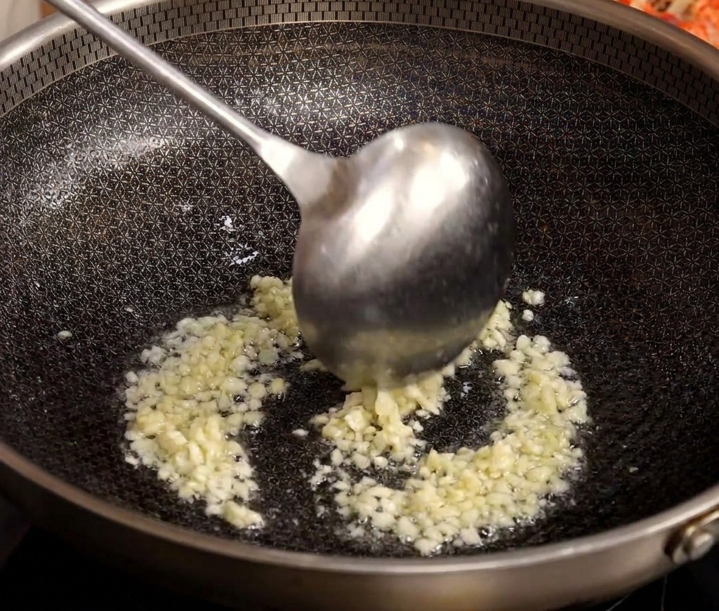 Un mestolo di metallo mescola laglio tritato mentre sfrigola e dora in un wok caldo con olio.