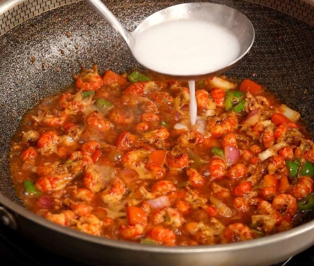 Un mestolo in acciaio inossidabile versa un composto bianco di amido al centro di una padella che sobbolle con gamberi rossi piccanti.