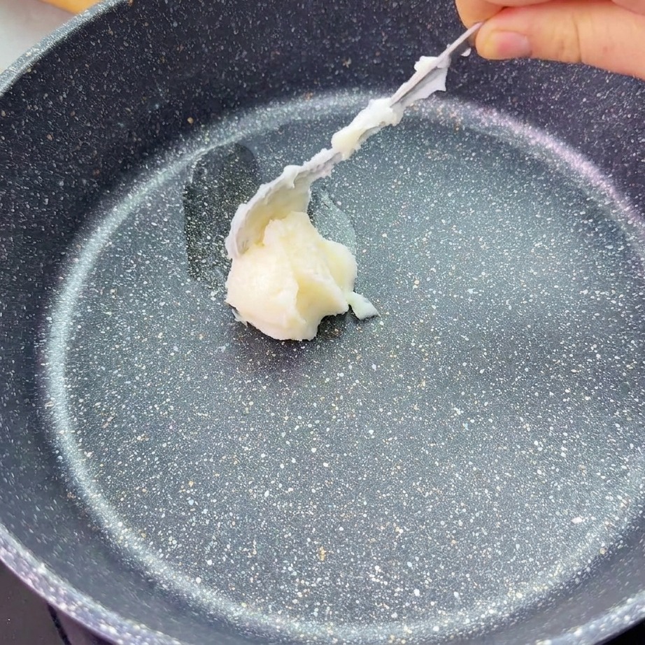 Un cucchiaio deposita una noce di strutto bianco al centro di una padella antiaderente scura.