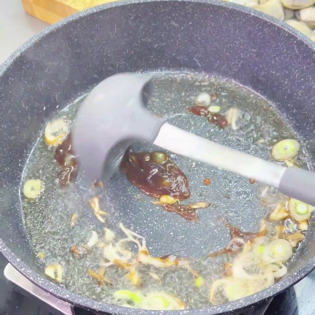 Salsa dostriche densa e scura mescolata ai cipollotti soffritti con una spatola in silicone grigia.