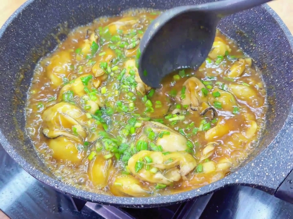 Un mestolo scuro mescola ostriche intere ed erba cipollina in una salsa bruna lucida e bollente.