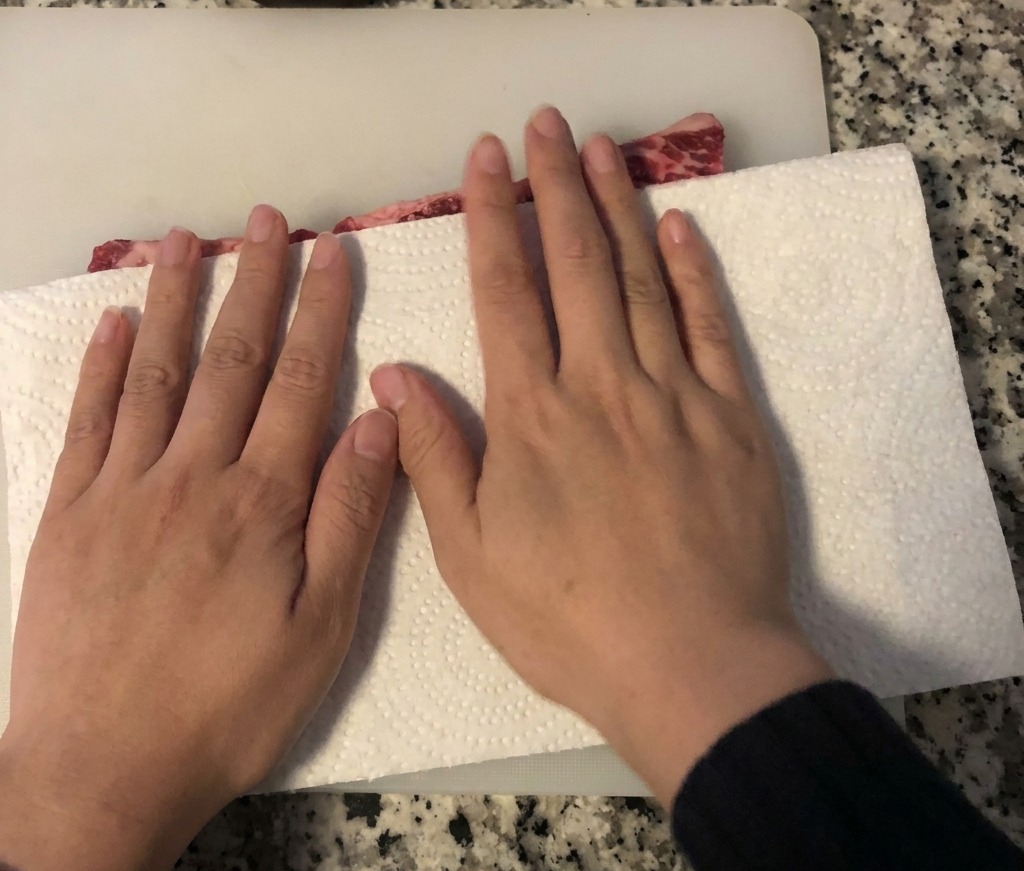 Mani che usano un tovagliolo di carta per assorbire lumidità dalle costolette di manzo crude disposte su un tagliere bianco.