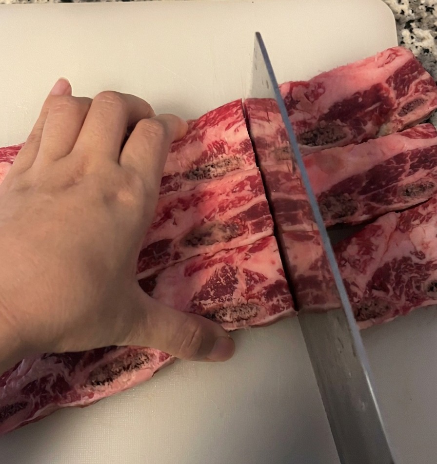 Una persona taglia un pezzo di costolette di manzo crude in singole porzioni con osso usando un grande coltello da chef su un tagliere bianco.