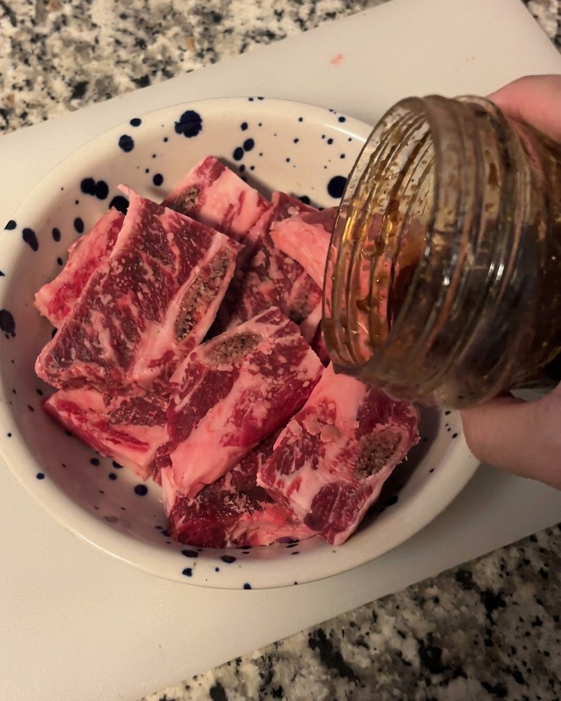 Salsa barbecue coreana scura e lucida versata da un barattolo di vetro su una pila di costolette di manzo crude affettate in una ciotola bianca a macchie blu.