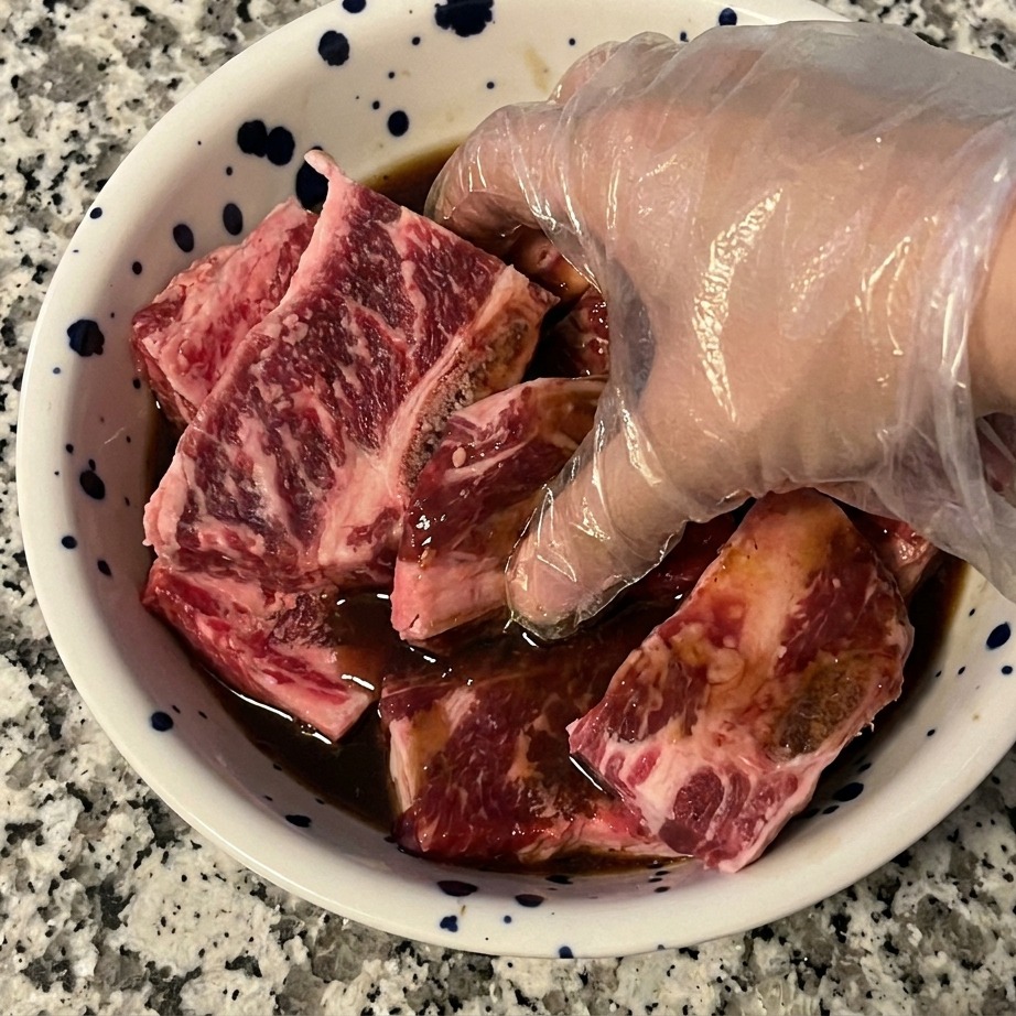 Una mano con un guanto di plastica trasparente mescola le costolette di manzo crude con una marinatura scura in una ciotola per garantire una copertura uniforme.