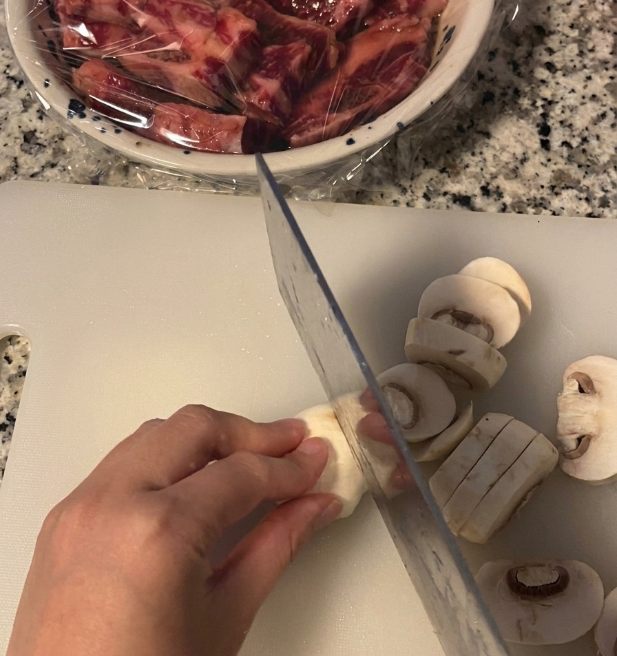 Una mano tiene un fungo champignon bianco mentre lo taglia a fette spesse con un coltello da cucina su un tagliere bianco.