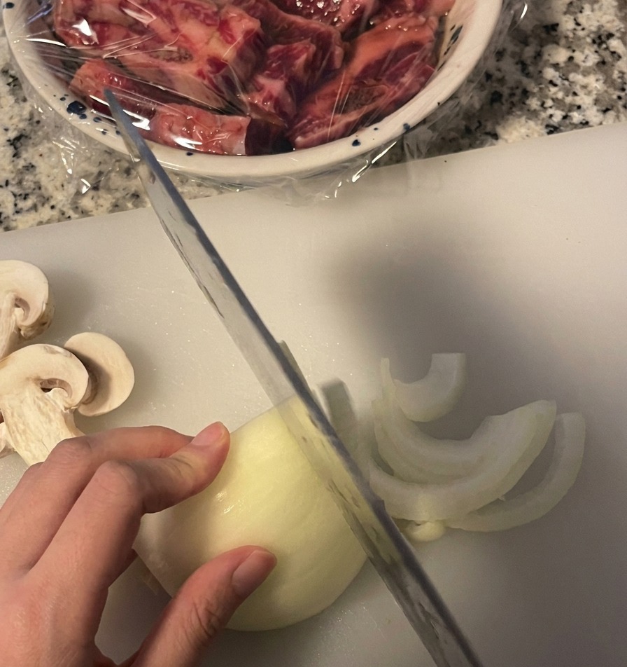 Una persona taglia una cipolla bianca a fettine sottili a mezzaluna su un tagliere bianco usando un coltello da cucina.