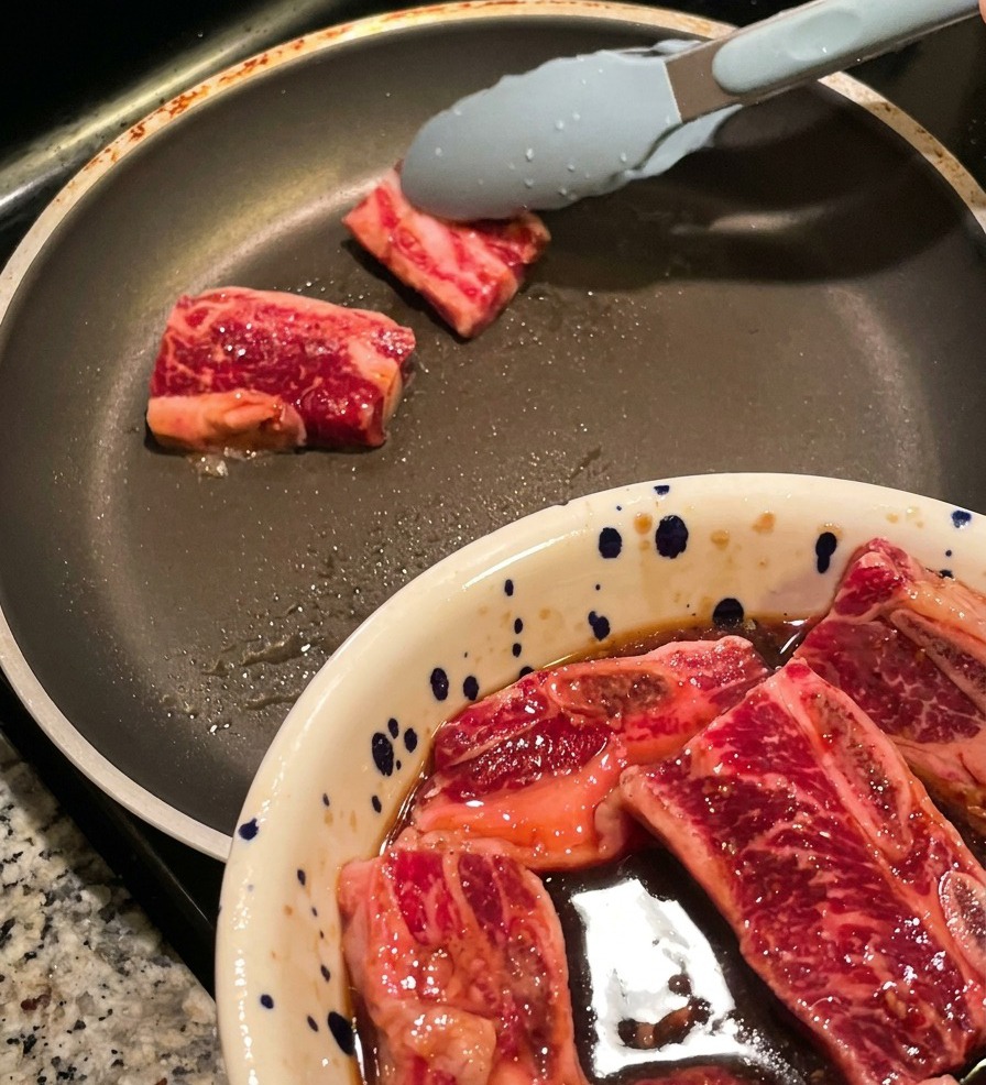 Pinze che posizionano spesse fette di costolette di manzo crude marinate in una padella calda, con una ciotola di altra carne visibile in primo piano.
