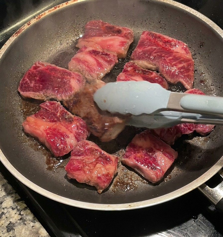 Uso di pinze con punta blu per girare le costolette di manzo rosolate in una padella, rivelando una crosta scura e caramellata sul lato cotto.