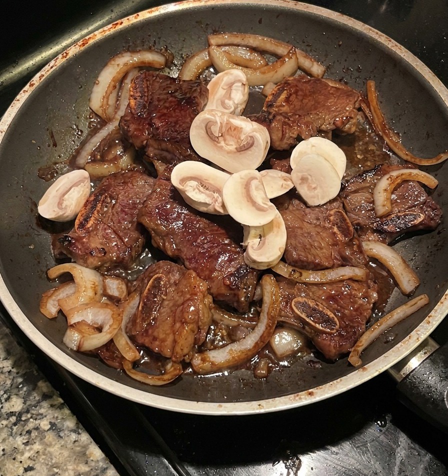 Funghi affettati saltati in padella con costolette di manzo e cipolle caramellate in una padella scura.