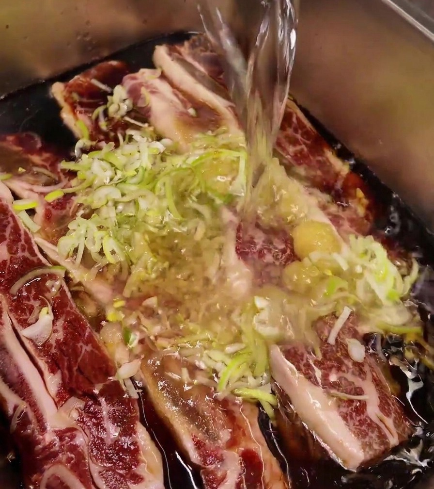 Getto dacqua versato in un vassoio dacciaio contenente costine di manzo, aglio e cipollotti.