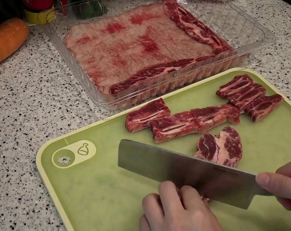 Una persona usa una mannaia per tagliare costine di manzo marmorizzate in piccoli pezzi rettangolari su un tagliere verde.