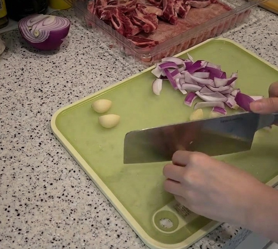 Strisce di cipolla rossa e spicchi daglio sbucciati disposti su un tagliere verde durante la preparazione.