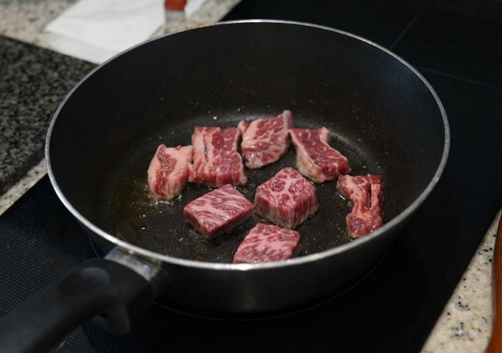 Pezzi di costine di manzo marmorizzate che rosolano in una padella scura, con i bordi che diventano bruni e opachi.