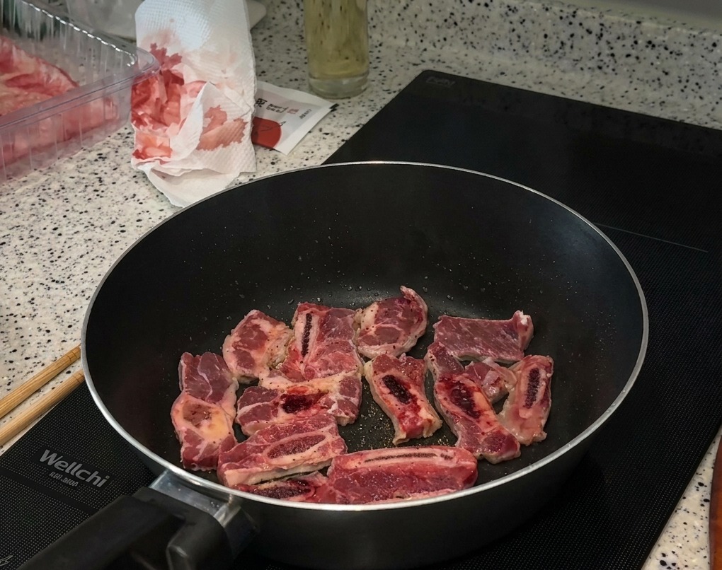 Pezzi di costine di manzo che rosolano in padella su un piano a induzione, con sale e pepe nero visibili sulla carne.