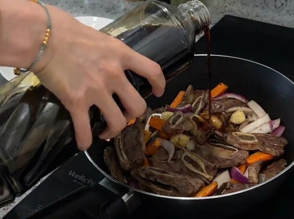 Primo piano di salsa di soia chiara versata da una bottiglia di vetro in una padella con manzo, carote e cipolle.